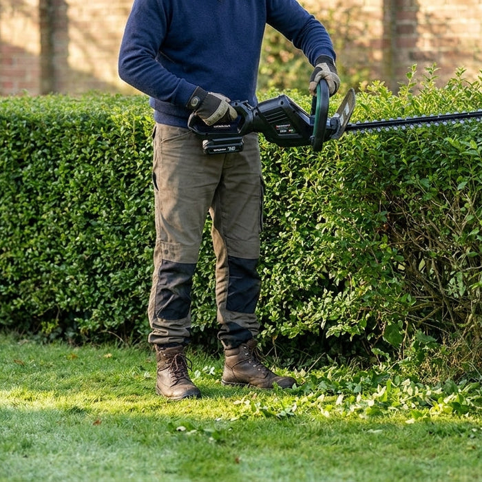 Gardener with a Spectrum cordless hedge trimmer working a British garden hedge in late spring